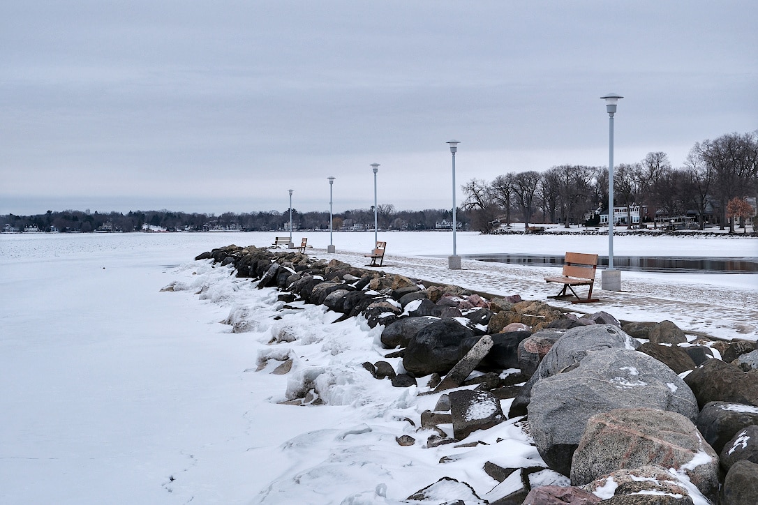 A concrete pier with large stones on a snow covered lake. Light poles and benches line the pier and trees are seen on the lake's distant shore.