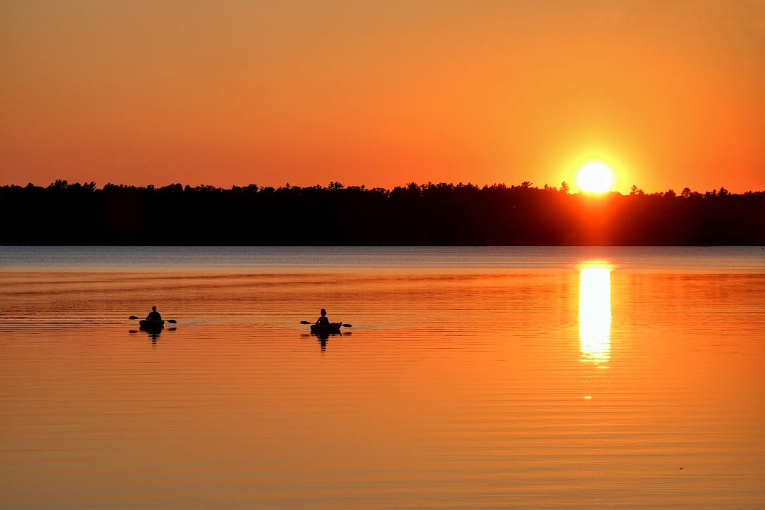 Two kayaers paddle on a lake as the sun sets over the distant shore. A deep orange sky is reflected in the calm water.