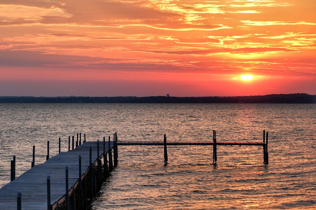 The sun setting though orange clouds over a lake. A pier leading into the lake is in the foreground.