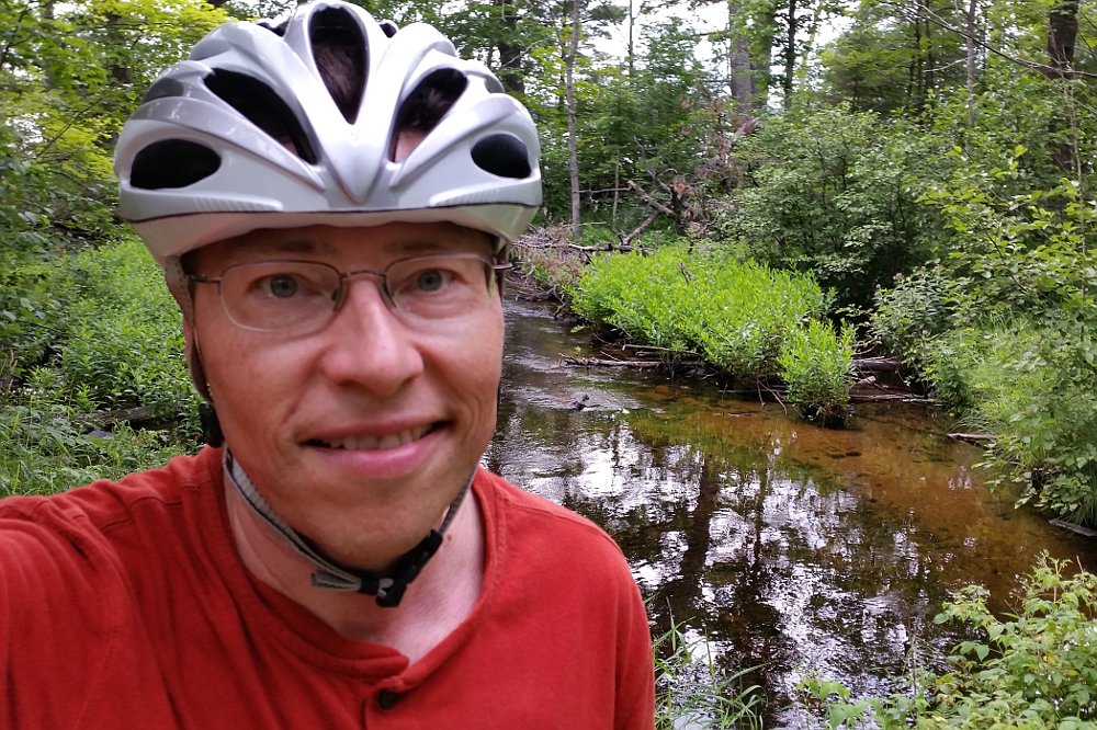 Self portrait of Jonathan Bloy wearing a bike helmet in front of a forest creek