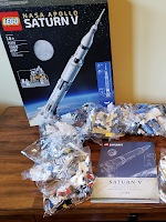 A Lego box for a Saturn V model and bags of Lego pieces surrounding an instruction manual on a desk