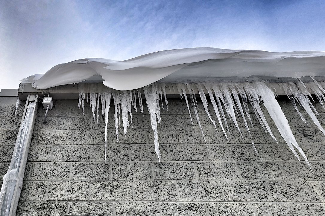 A curvy snowdrift and icicles hang over the edge of the roof, along a grey brick wall.