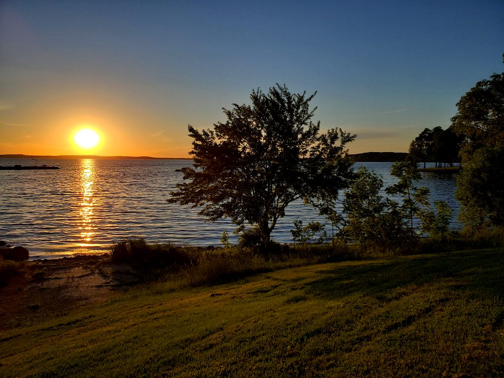 Sun just at the edge of the horizon on a lake, with trees and grass in the foreground