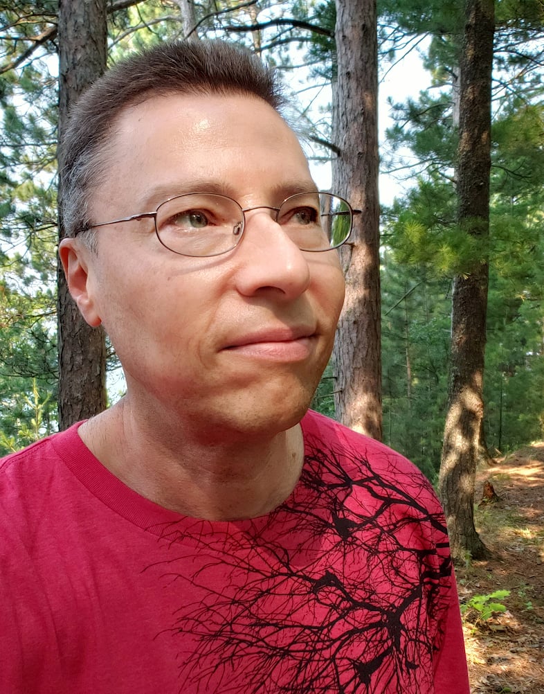 Jonathan Bloy standing in a forest, looking thoughtfully toward the pine trees