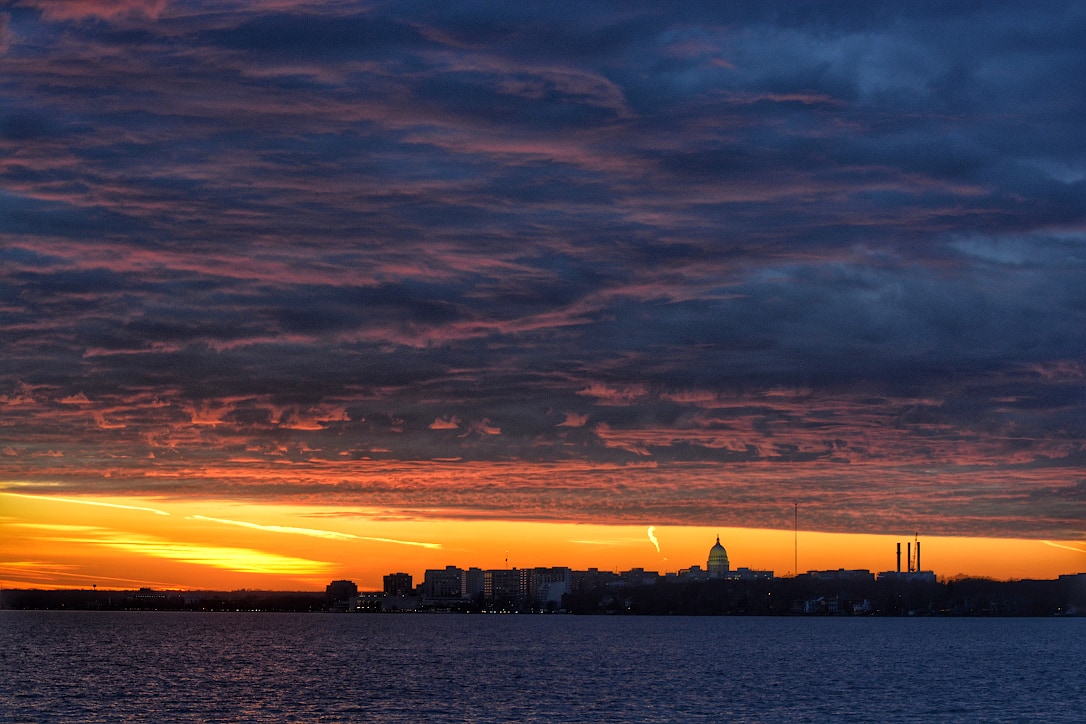 Purple and blue clouds above an orange colored band of sky.  The downtown Madison skyline can be seen from across a lake.