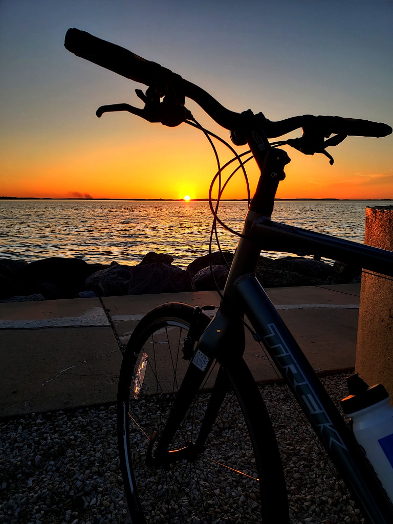 The sun sets just above the horizon of a lake, behind the handlebars and front wheel of a bicycle.