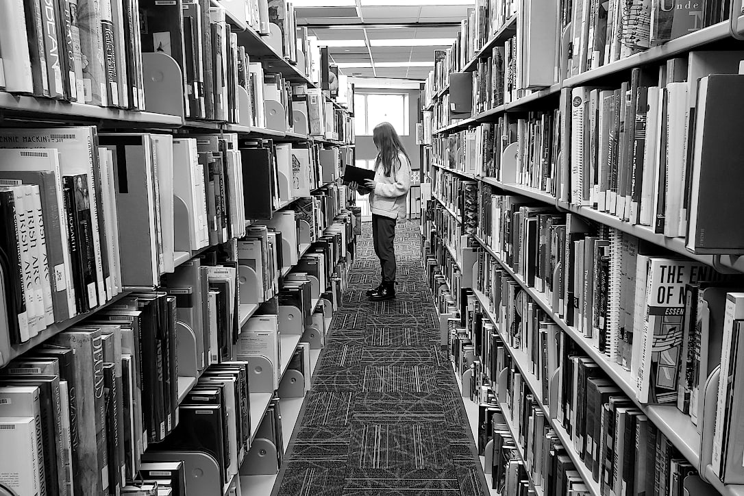 A student stands in the middle of a row of library bookshelves, looking at a book.