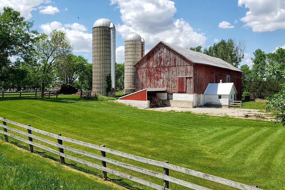 A red barn with two silos behind a freshly mowed lawn. A wooden fence is in the foreground.