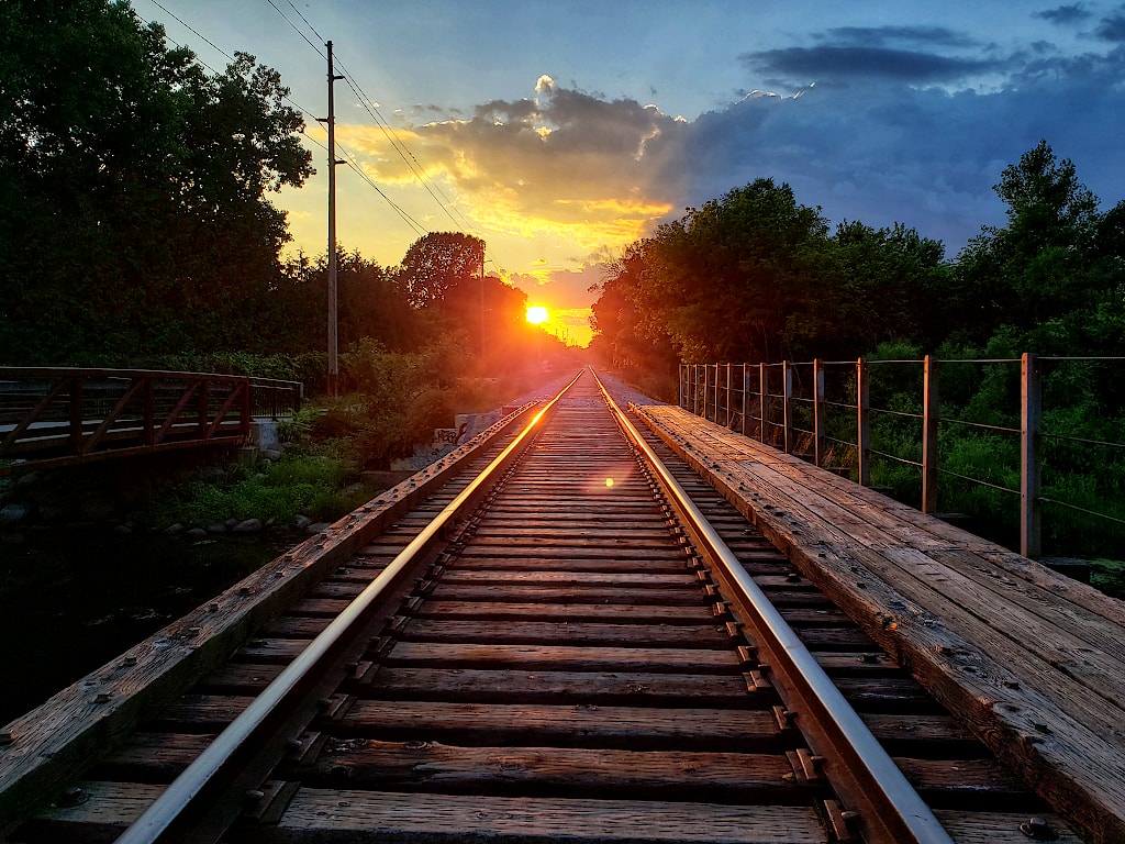 Railroad tracks leading off into the distance reflecting the late-day sun. In the background are trees, clouds in the sky, and the sun, which is low in the sky.