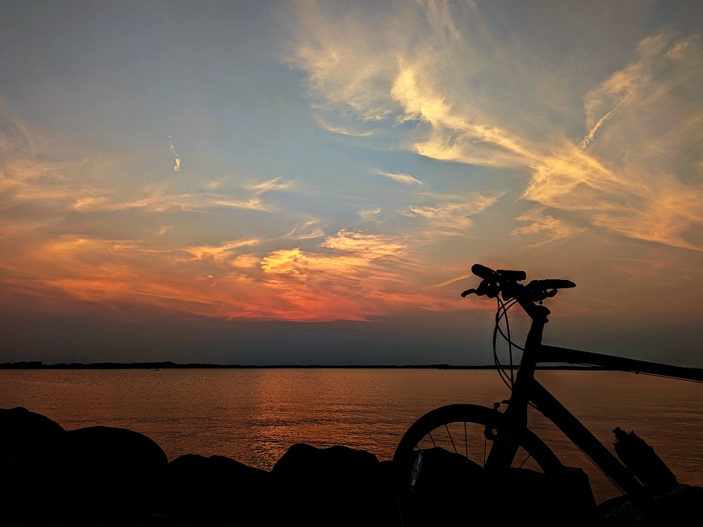 Silhouette of a bicycle and shore rocks, in front of an orange, yellow and hazy blue sky.