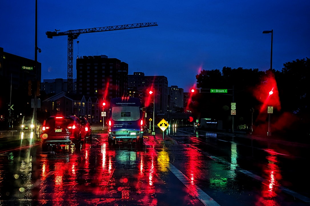 Traffic signals and brake lights from stopped vehicles are reflected in the wet pavement. Silhouettes of buildings and a crane are in the background under a twilight blue sky.