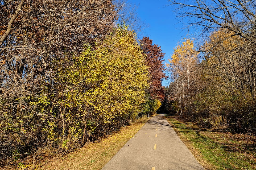 A paved path leads into the woods.  On either side of the trail are trees with some late-season color.