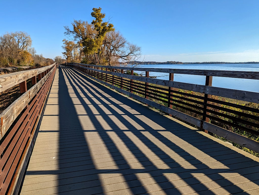 View of a long boardwalk extending into the distance with shadows from its railings. A large tree is in the distance and a lake on the side.
