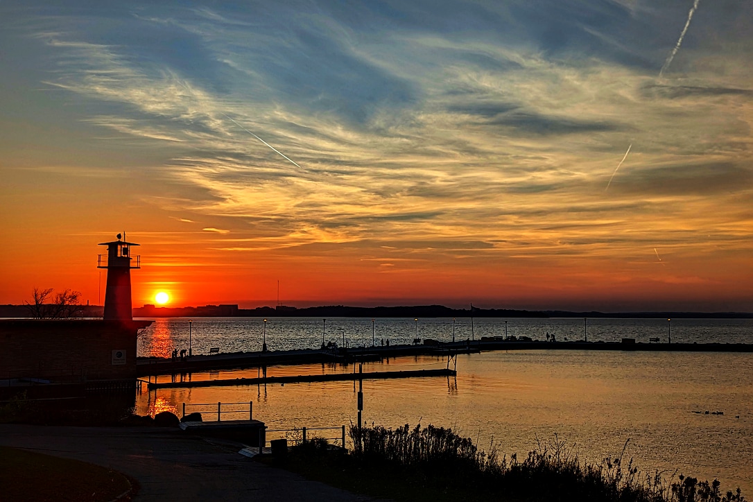 The sun just over the horizon, next to a light house and pier. The sky has wispy red, orange, and white clouds.
