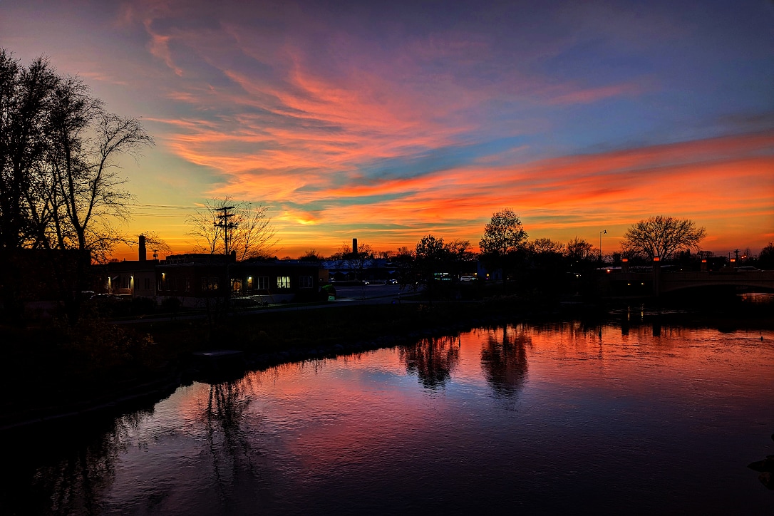 A red and blue sky with touches of orange is reflected in a river.  Along the shore are trees, buildings, and a utility pole in silhouette.
