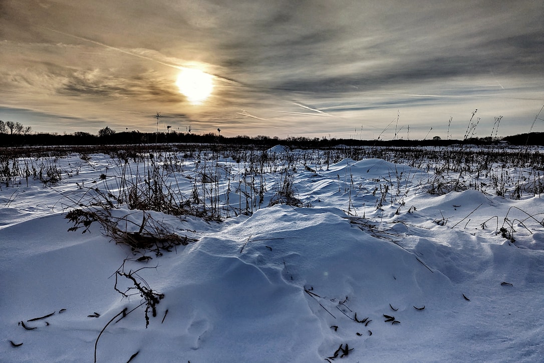 The sun shines through streaky clouds, over a prairie covered with snow drifts. Scattered dried plants poke up through the snow.