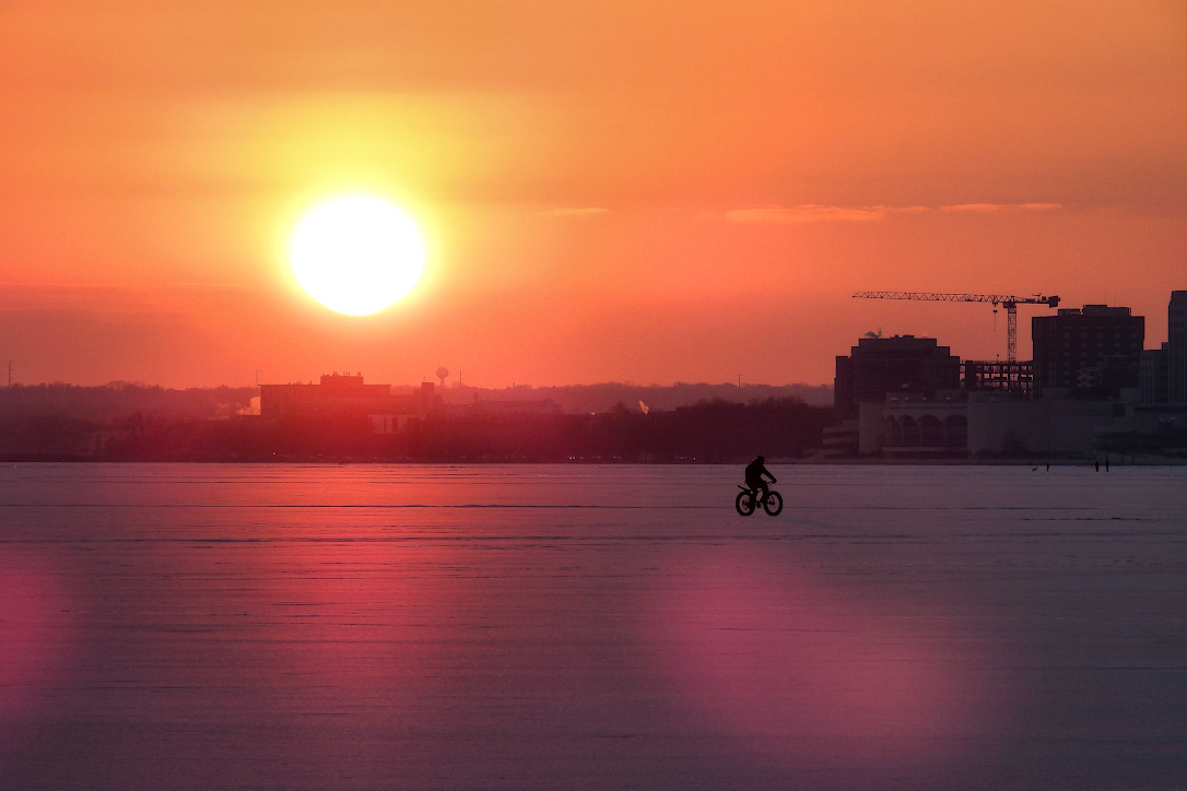 Sun shining in an orange sky, just above the horizon.  A bicyclist is riding across a snow covered, frozen lake.