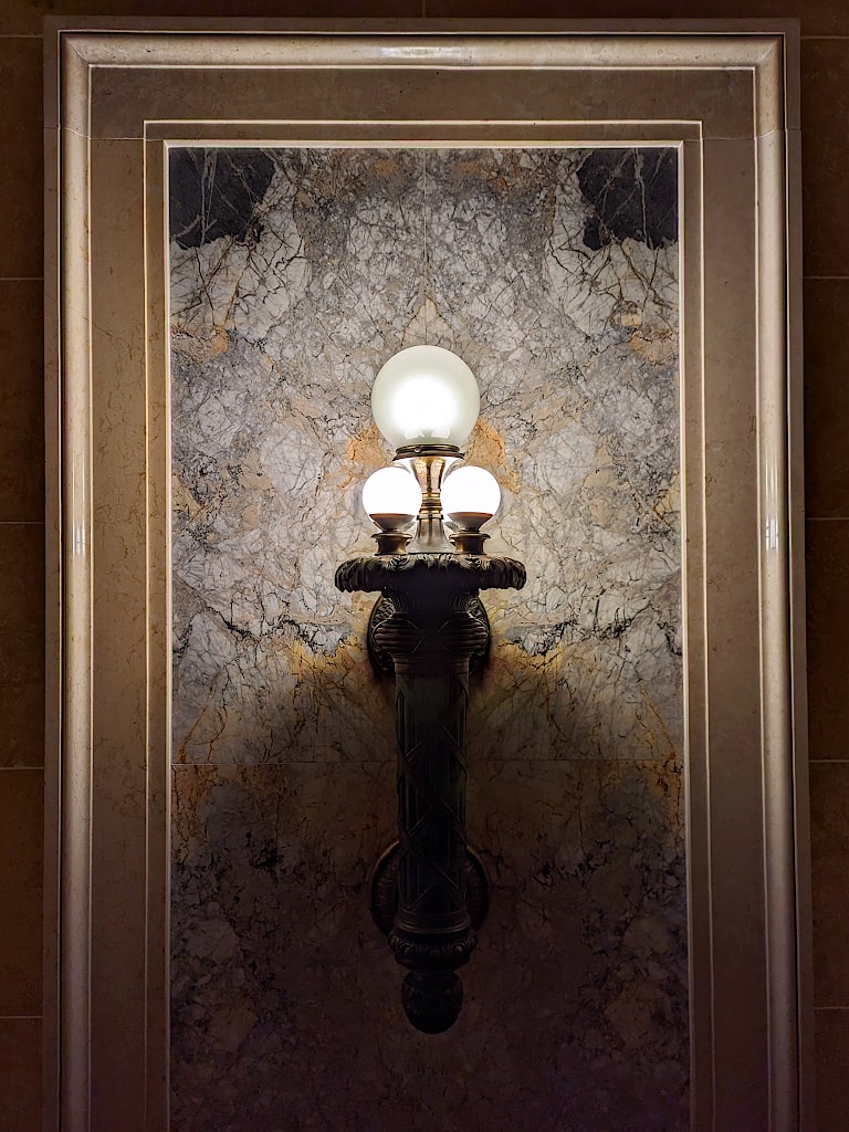 An ornate wall sconce. Behind it are strongly veined marble tiles framed with molding.