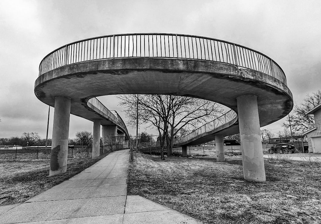 A sidewalk leads to a ramp for a pedestrian overpass. The fenced ramp takes a loop in the foreground supported by two large pillars, and leads off in the distance over the highway.
