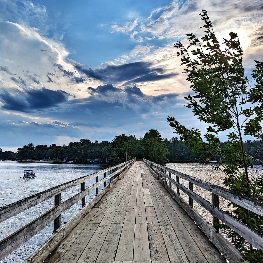 View from the top of a wooden trestle leading across a lake. Streaky clouds with blue sky are directly overhead and storm clouds are approaching from the distance.