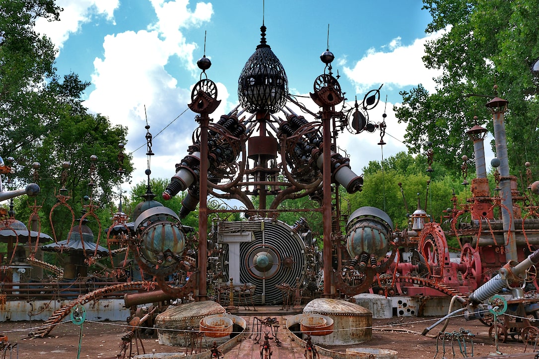 Large sculpture made of scrap metal pieces. A tall tower with an egg shaped cage is in the center. Two other tall structures are next to the tower, surrounded by many other large and small pieces.