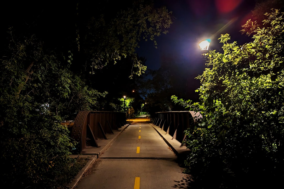 A lamppost lights up a paved bike path, over a former railroad bridge. The path continues into the distance.