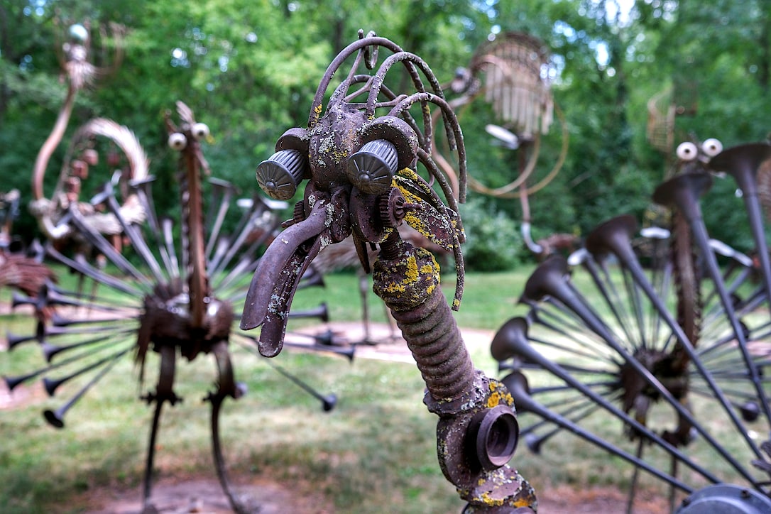 Closeup of a bird's head made out of various scrap metal pieces. In the background, out-of-focus, are other bird-like creatures.