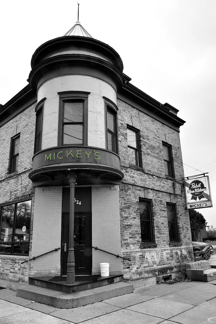 An old brick, two-story building. The corner of the structure has a door with a turret above it on the second floor.  Above the door is a green neon sign that says Mickey's.