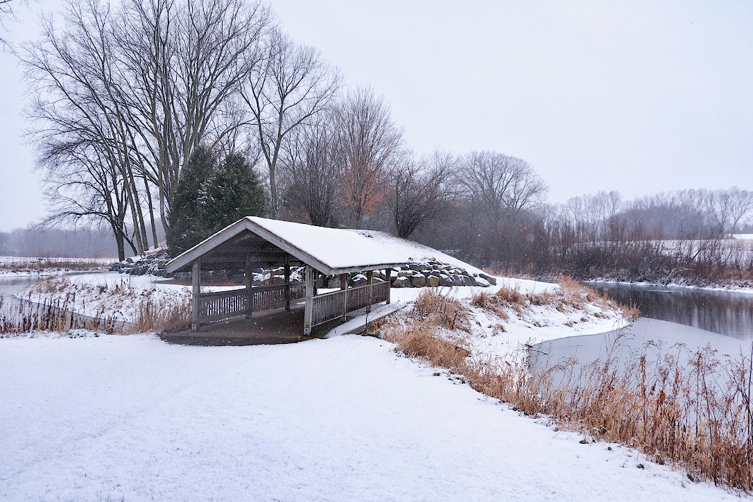 An area of freshly fallen snow in front of a covered footbridge that crosses a pond. Prairie grass and trees line the shore of the pond.