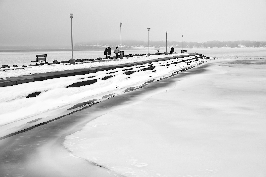 A concrete pier juts out into a frozen lake on a cloudy and slightly foggy day. A few people are walking along the pier.